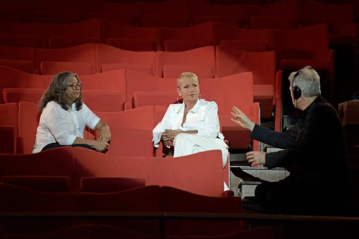Marlene Mattos e Xuxa Meneghel sentadas em uma plateia de teatro, ambas de roupa branca, sendo dirigidas para documentário