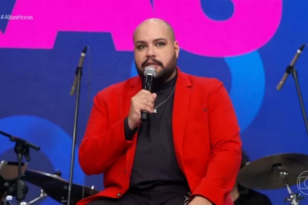 Tiago Abravanel de blazer vermelho e camisa de gola alta preta, segurando microfone, sentado