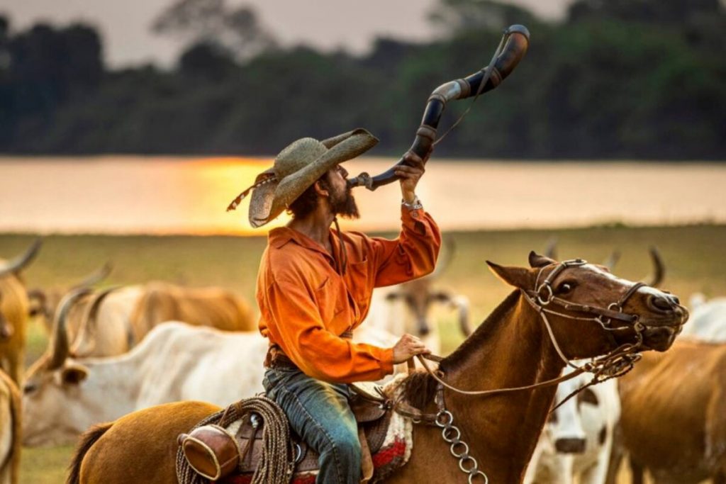 Irandhir Santos montado no cavalo, de calça jeans e camisa laranja, tocando berrante