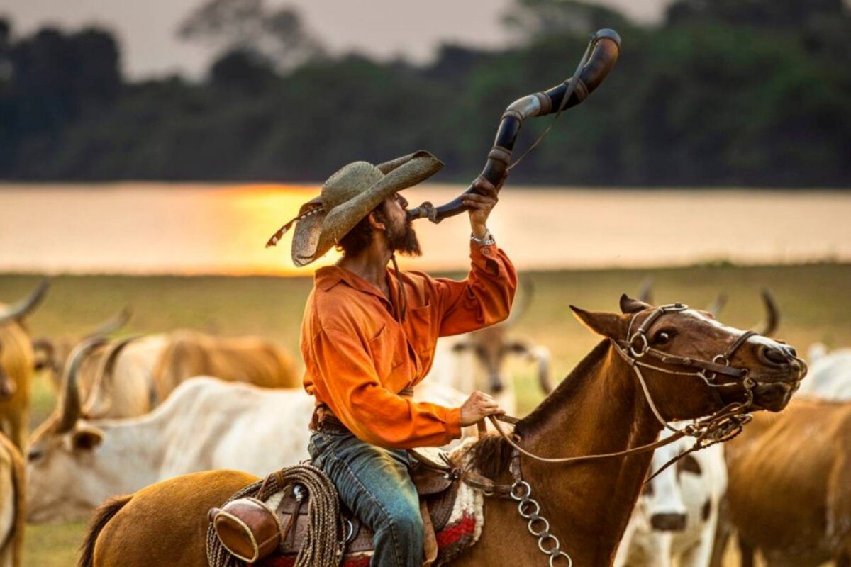 Irandhir Santos montado no cavalo, de calça jeans e camisa laranja, tocando berrante