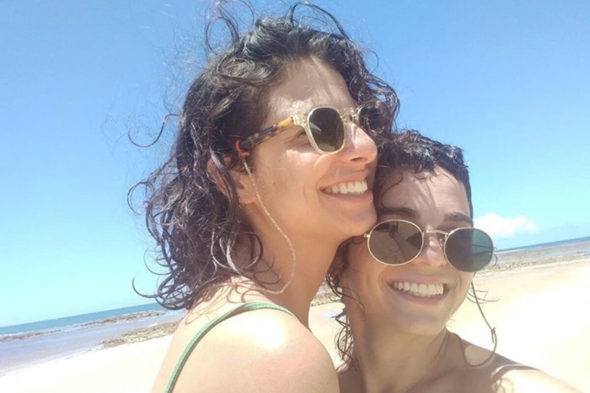 Júlia Anquier e o ator trans Flow na praia, de óculos escuros, sorrindo