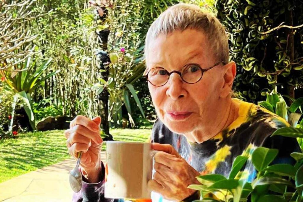 Rita Lee segurando uma caneca, de cabelos curtos e camisa estampada, no jardim