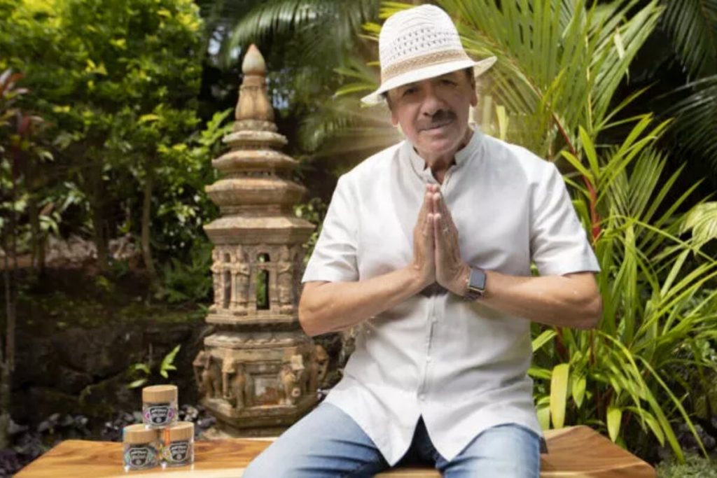 Carlos Santana de chapéu branco, calça jeans, camisa branca, sentado com as mãos juntas 