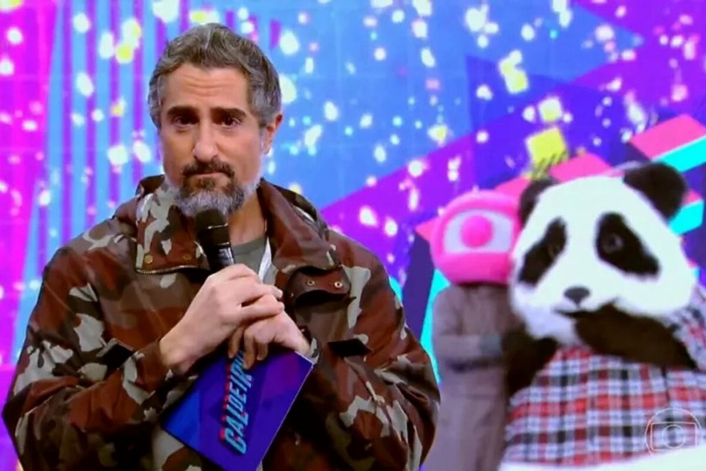 Marcos Mion emocionado, de casaco camuflado, segurando microfone, no palco do 'Caldeirão'