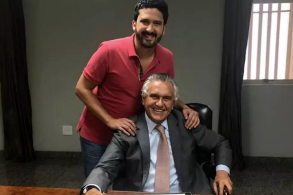 Ronaldo Caiado Filho, em pé, de camisa vermelha e calça jeans, abraçado ao pai, Ronaldo Caiado, sentado, de terno