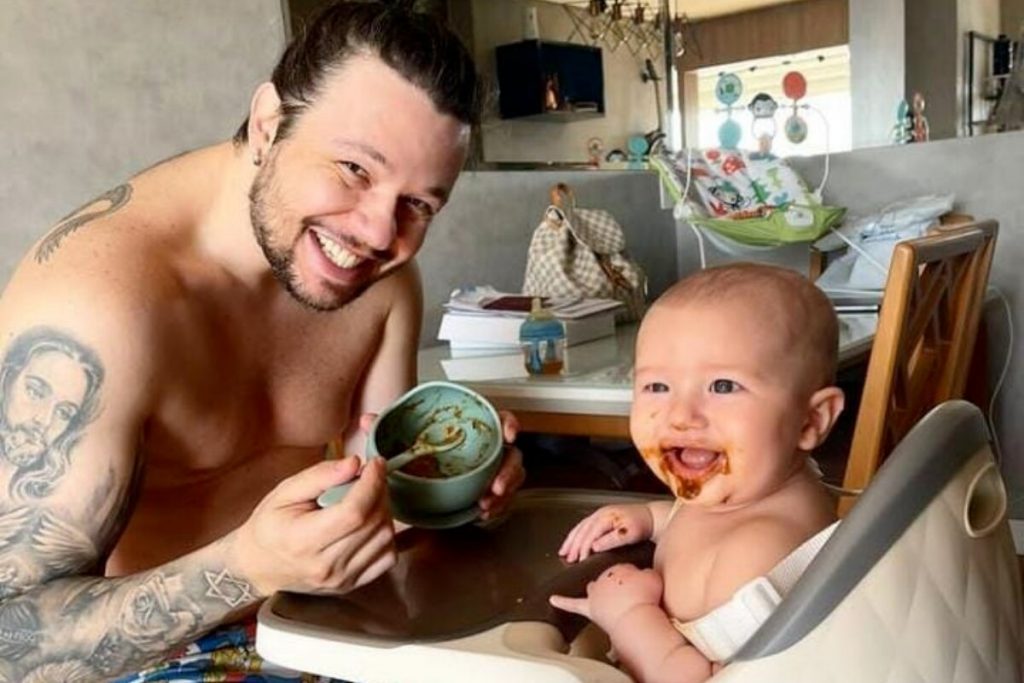 Bruno KLB sem camisa, sorrindo, dando papinha para o filho, que está sentado na cadeirinha