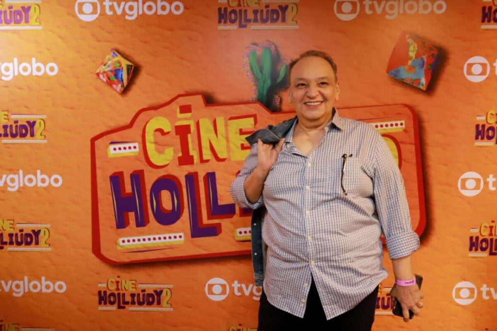 Cacá Carvalho de camisa social cinza claro e calça preta, posado diante do backdrop de fundo laranja