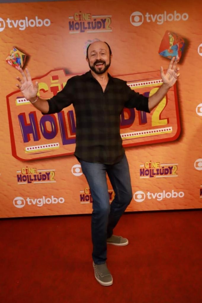 Carri Costa de calça jeans e camisa xadrez marrom, fazendo pose diante do backdrop laranja