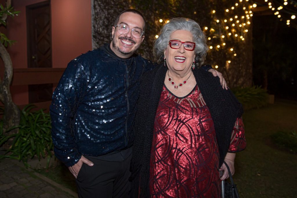 Amigo de Mamma Bruscchetta de casaco azul escuro, abraçado a ela, que está de vestido vermelho brilhoso e casaco preto, apoiada em uma bengala