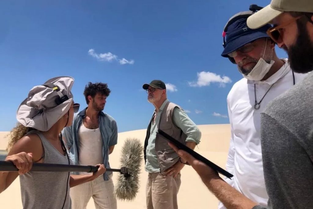 Chay Suede e Marcos Caruso, ambos de bermuda behe e camisa, gravando cen de Travessia nos lençóis maranhenses, com assistemtes segurando microfones e diretor de cena