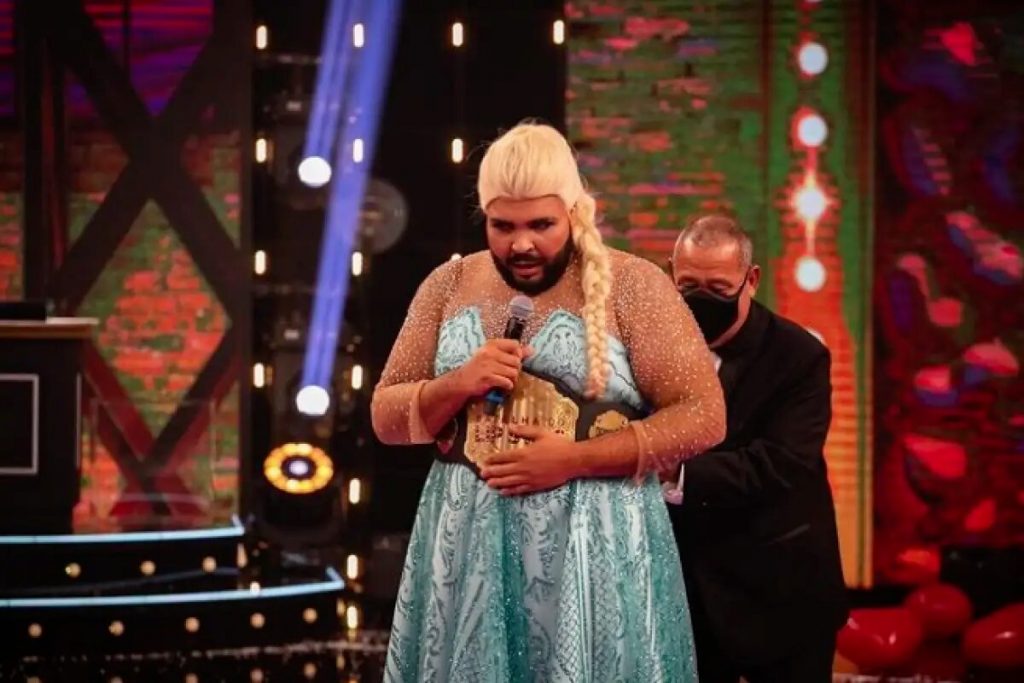 Homem de preto tentando colocar um cinturão em Paulo Vieira, que está de vestido azul e peruca loira, como a Frozen