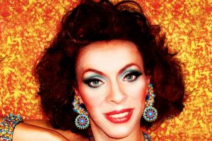 Retrato colorido de Cláudia Celeste, com brincos grandes, fundo laranja