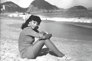 Cláudia Celeste sentada na areia da praia, de roupa normal 