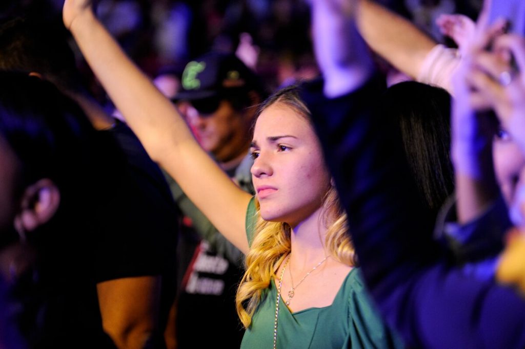 Jovem loira com o braço estendido, para a frente, de blusa verde