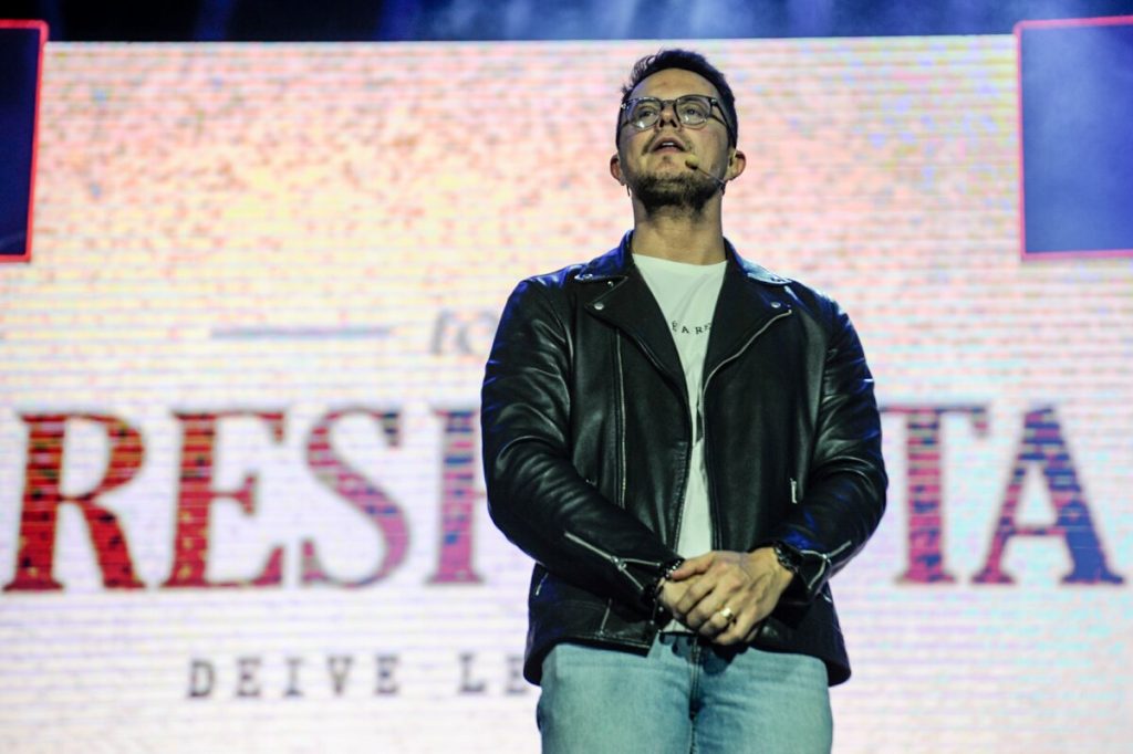 Deive Leonardo de jaqueta preta e camisa branca, de óculos de grau, com as mãos unidas na frente do corpo