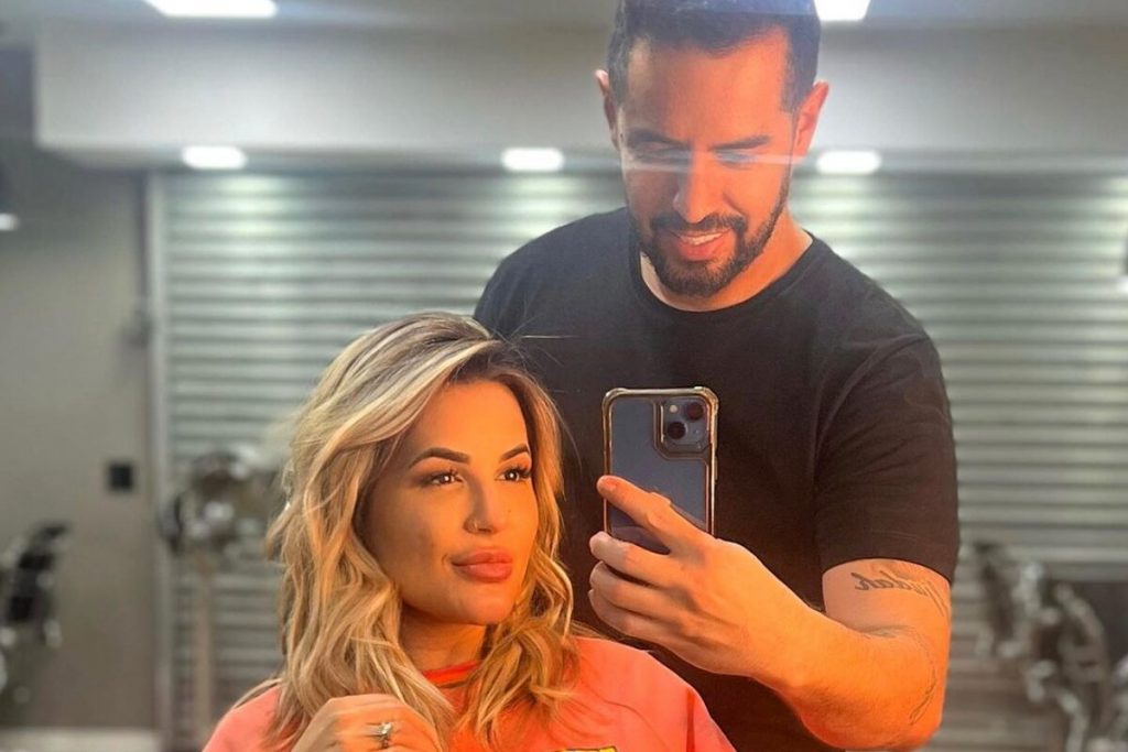 Deolane Bezerra de moletom laranja, sentada, co os cabelos produzidos pelo hair stylist, de camisa preta, que faz selfie dos dois