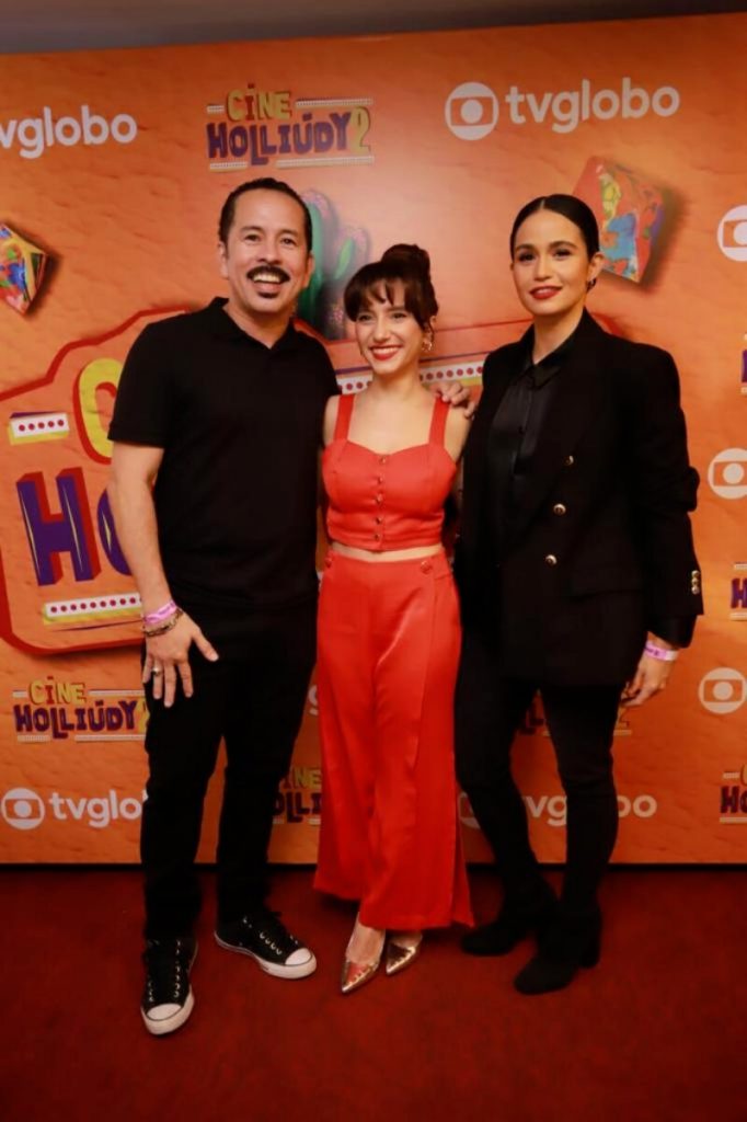 Edmilson Filho, todo de preto, Luisa Arraes, de calça e cropped vermelho, e Nanda Costa, toda de preto, abraçados diante do backdrop de fundo laranja