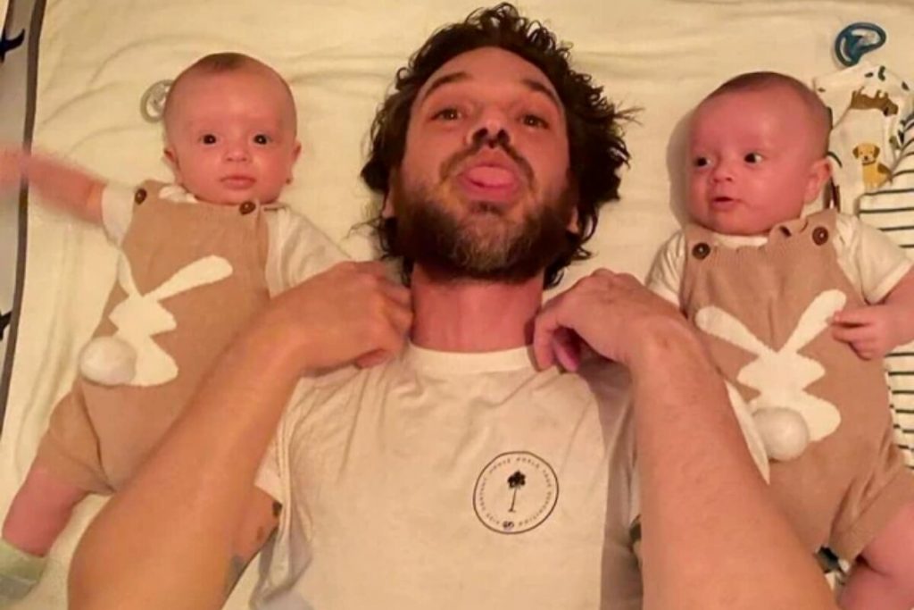 Emílio Dantas e os filhos gêmeos, deitados na cama, o ator fazendo careta, de camisa branca, segurando a mão de cada um deles