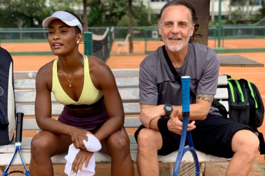 Erika Januza e Gustavo Novaes, em cena de Amor de Mãe, sentados, ela de top amarelo, viseira branca e bermuda preta, segurando uma toalha, ele segurando uma raquete de tênis  