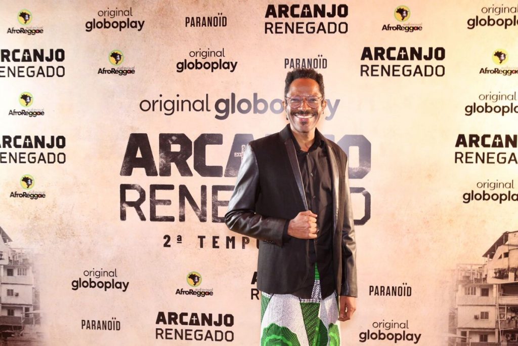Flávio Bauraqui de saia verde e branca e jaqueta preta de couro, posado no backdrop de Arcanjo Renegado