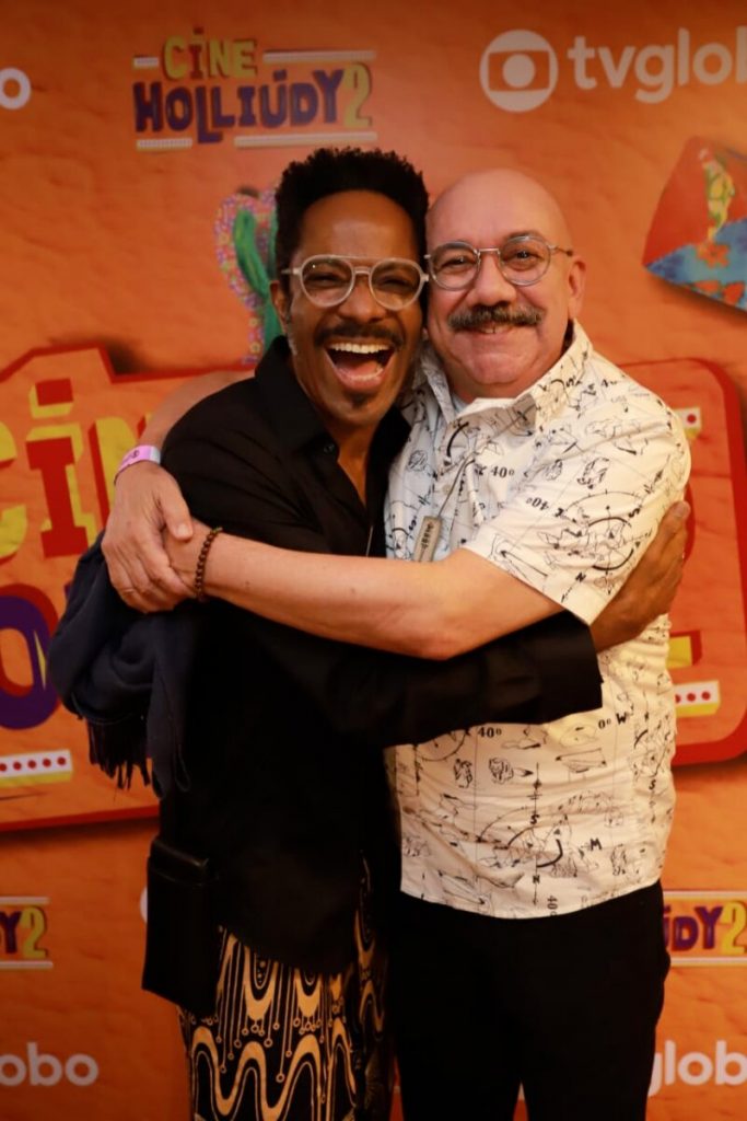 Flavio Bauraqui de camisa preta, abraçado a Frank Menezes, de camisabranca estampada, ambos sorrindo diante do backdrop de fundo laranja