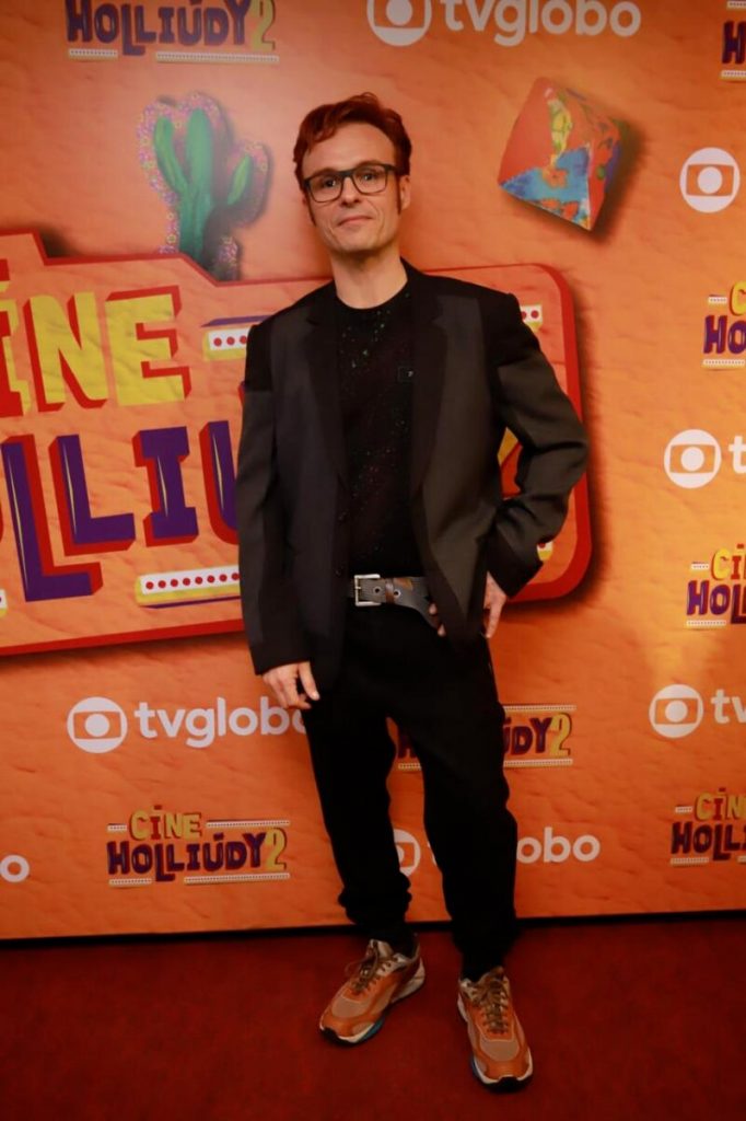 Gustavo Falcão de calca, camisa e blazer preto, posando no backdrop da série, que tem a logo no fundo laranja