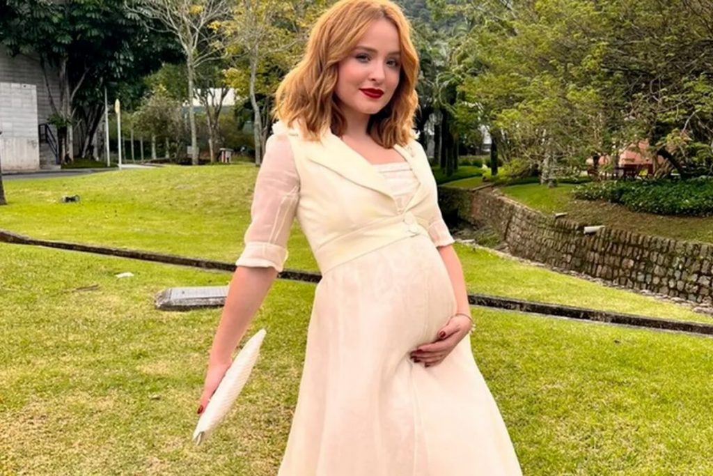 Larissa Manoela de vestido amarelo claro, segurando uma bolsa de mão, com barriga cenográfica, na fachada do estúdio de Além da Ilusão