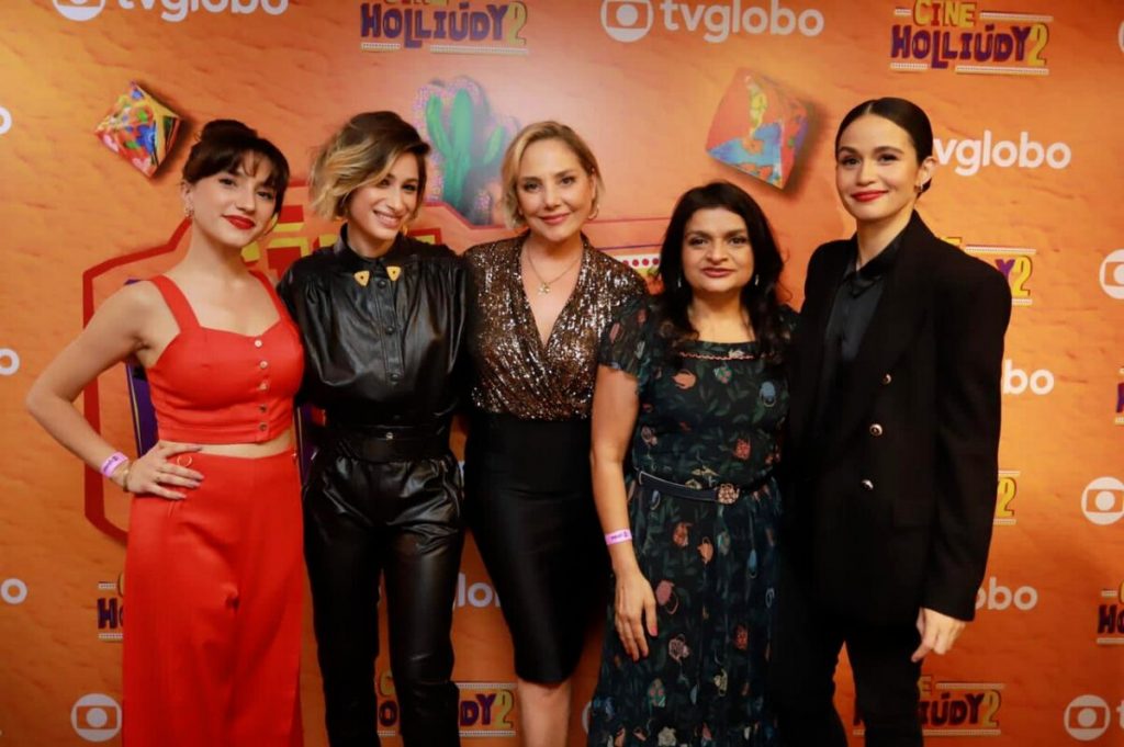 Lorena Comparato de calça e cropped vermelho, Luisa arraes de calça e blusa preta. Heloisa Perissé de blusa doirada e saia preta, Solange Teixeira de vestido estampado e Nanda costa de calça e blazer preto, de cabelo rpeso, abraçadas diante do backdrop de fundo laranja