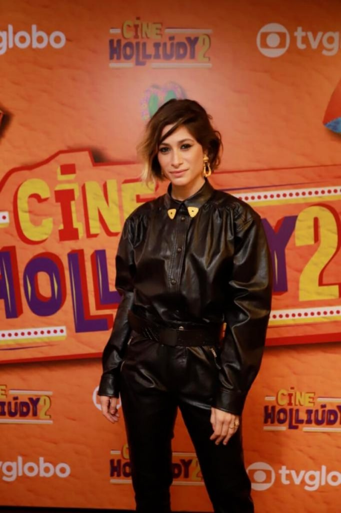 Luisa Arraes toda de preto, posando no backdrop da série, que tem a logo no fundo laranja