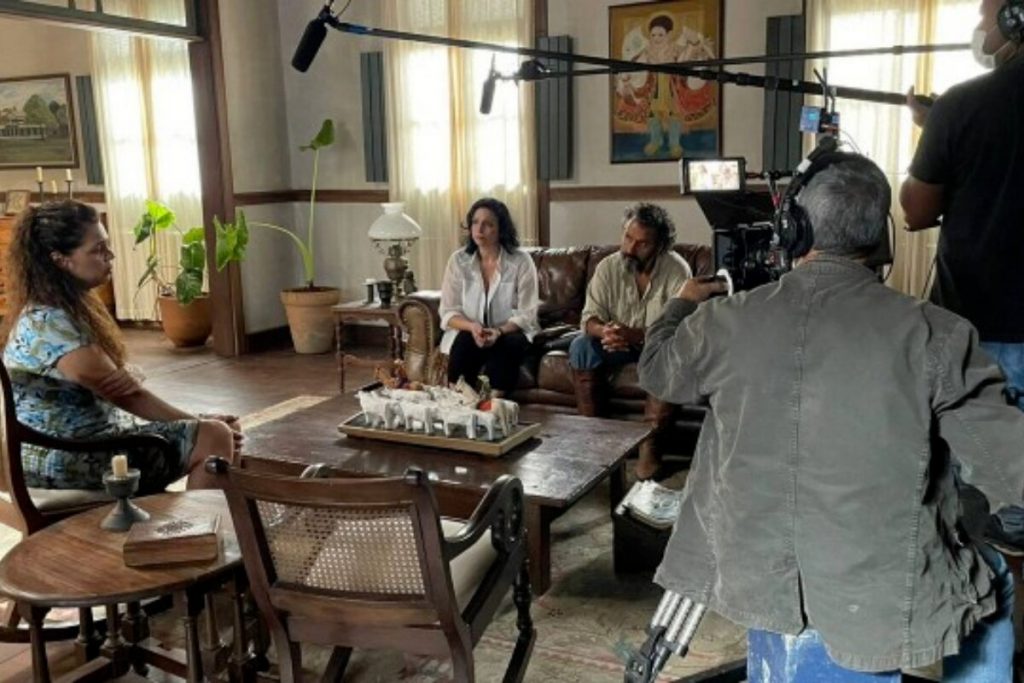 Bastidores da gravação de Pantanal com Maria Bruaca, José Leôncio e a advogada, sentados na sala, com câmera filmando e assistente segurando microfone