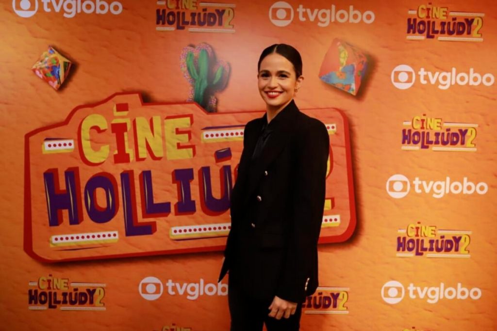Nanda Costa de cabelo preto, calçã e bçazer preto, posando no backdrop da série, que tem a logo no fundo laranja