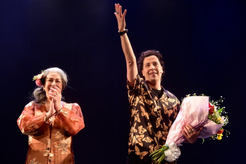 Nívea Maria aplaudindo e Arlindo Lopes segurando buquê de flores, saudando o público