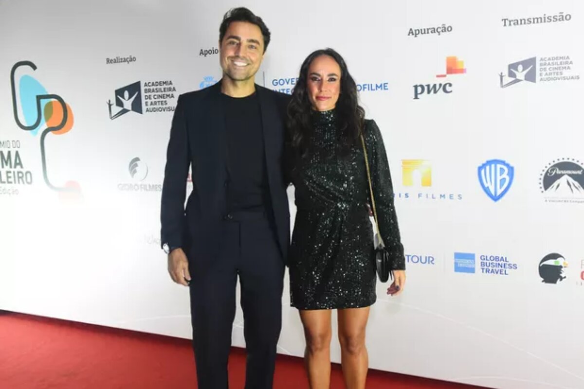 Ricardo Pereira e Francisca Pinto, ambos de preto, ela de vestido ele de blazer, calça e camisa social, sorrindo, abraçados, no tapete vermelho do Prêmio de cinema Brasileiro