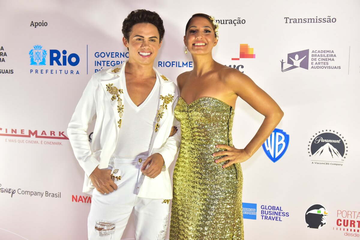 Silvero Pereira de branco com detalhes dourados e Camila Pitanga de vestido dourado e cabelos presos, com a mão na cintura