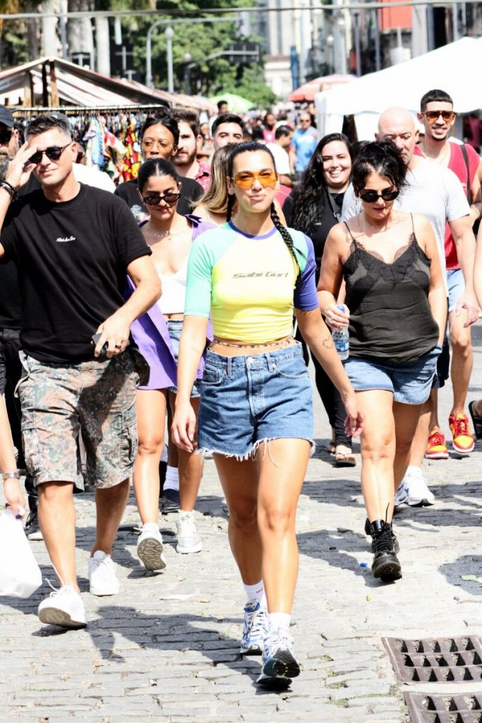 Dua Lipa com camisa verde e amarela, bermuda jeans, cercada por seguranças, andando na Feira do Lavradio, na Lapa, RJ