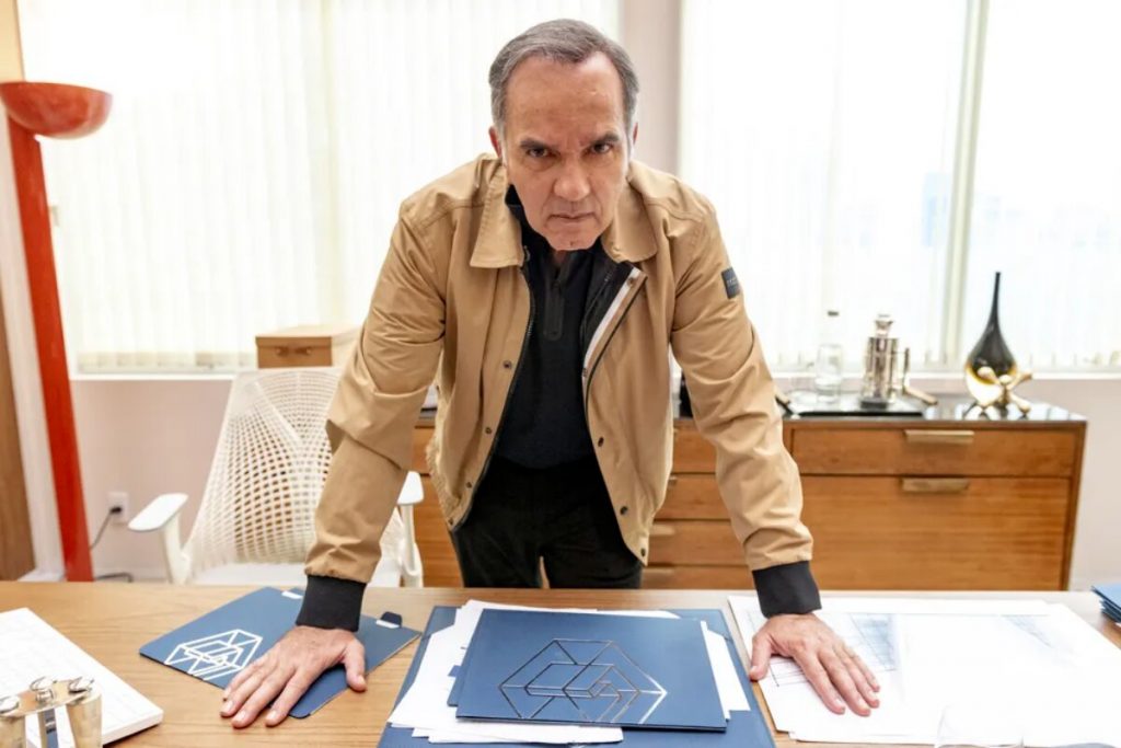 Humberto Martins caracterizado como Guerra,em Travessia, debruçado sobre uma mesa de escritório, de jaqueta bege e camisa preta