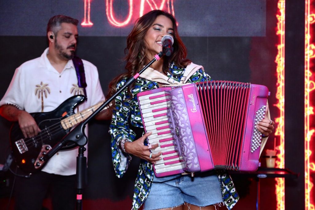 Lucy Alves de short jeans, blusão estampado verde e branco, cantando e tocando sanfona cor de rosa