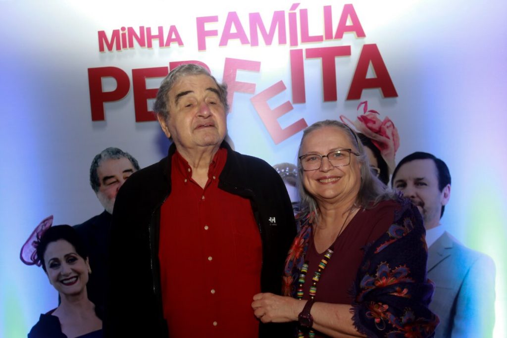 Otavio Augusto de camisa vermelha e casaco preto, de braços dados com a mulher, Cristina Mullins, de vestido estampado