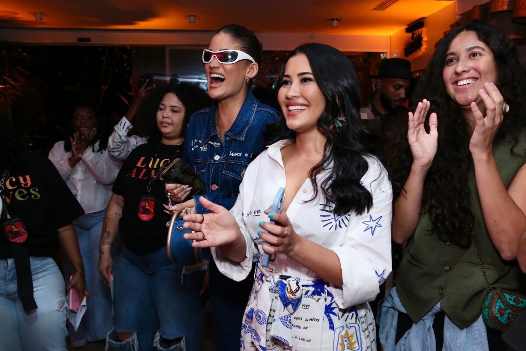 Thaynara OG de branco estampado ao lado de Laura Fernandez, que veste jeans e óculos escuros, ambas assistindo show