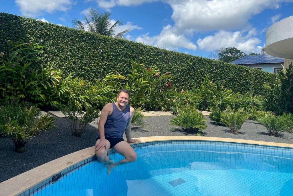 Ximbinha sentado na borda da piscina, de camiseta azul e sunga preta 