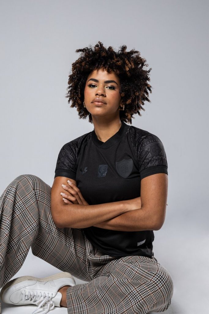 Modelo negra com camisa preta temática da Consciência negra, sentada no chão, de braços cruzados, calça xadrez cinza e marrom