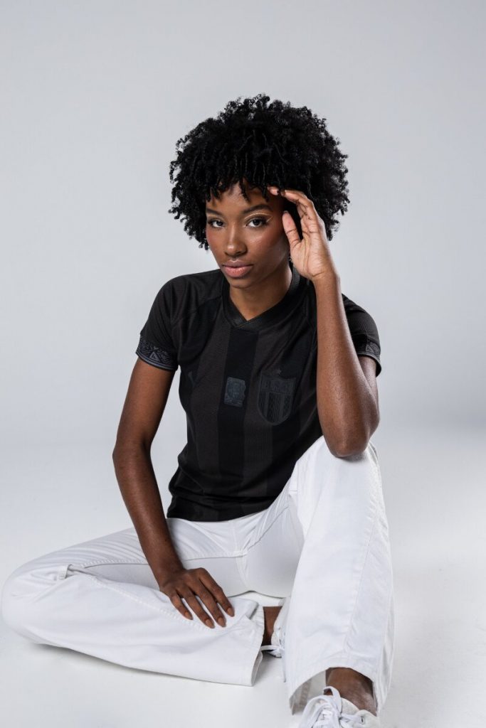 Modelo negra com camisa preta temática da Consciência negra, sentada no chão, com o braço esquerdo no cabelo e o outro sob a perna, de calça branca