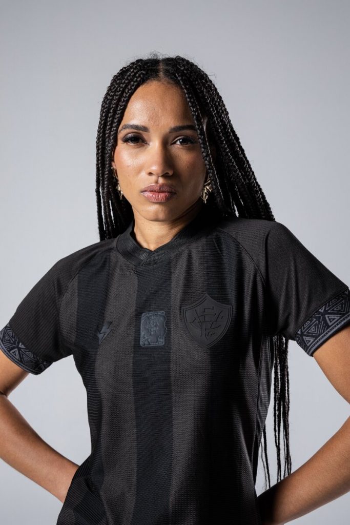 Modelo negra com camisa preta temática da Consciência negra, em pé com as mãos na cintura, d etranças rastafari