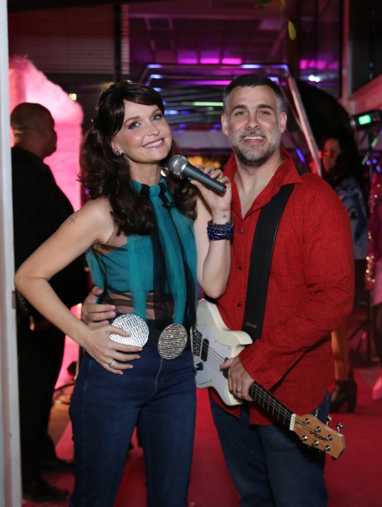 Fernanda Rodrigues de calça jeans, camisa verde, segurando microfone,  e Raoni Carneiro com uma guitarra, de camisa vermelha e calça jeans
