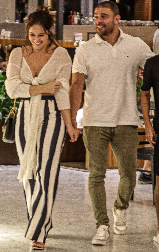 Diogo Nogueira de mãos dadas com  Paolla Oliveira, andando no corredor do shopping, ela de calça de listras pretas e brancas, ele de calça marrom e camisa branca