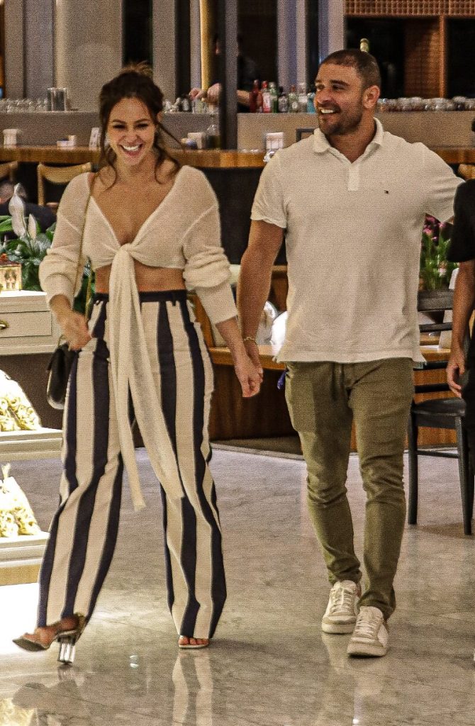 Diogo Nogueira de mãos dadas com  Paolla Oliveira, andando no corredor do shopping, ela de calça de listras pretas e brancas, ele de calça marrom e camisa branca