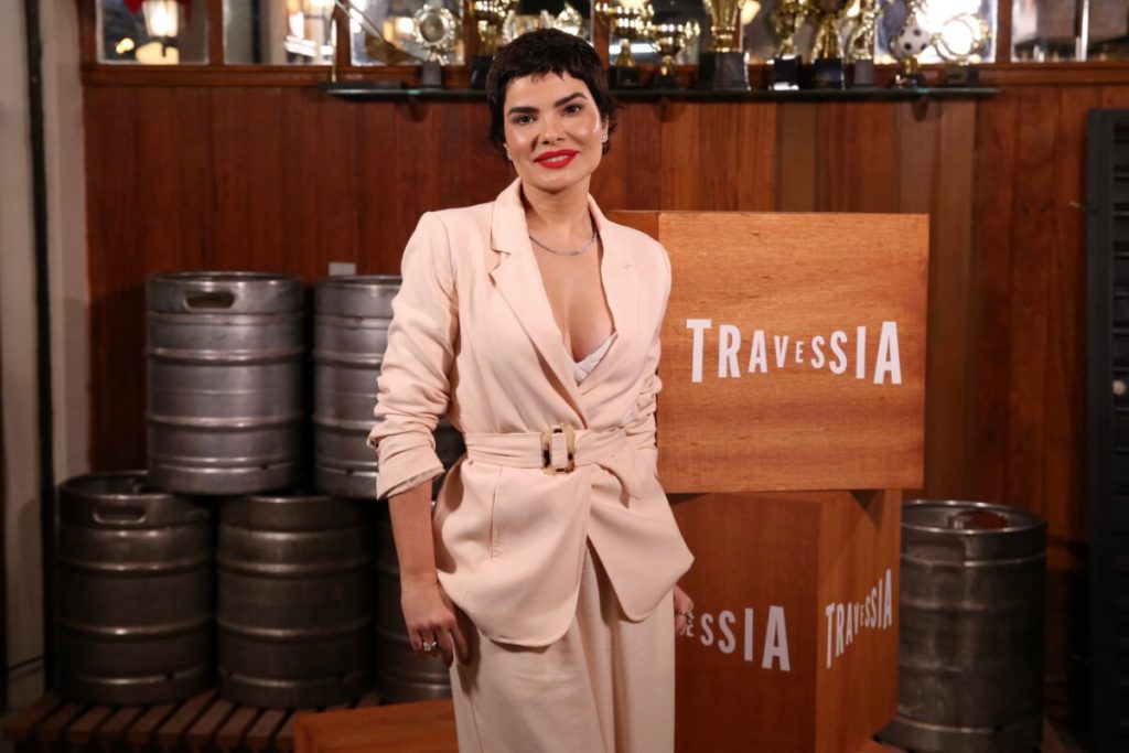 Vanessa Giácomo com os cabelos curtos, blazer e calça bege, no cenário do bar de 'Travessia', sorrindo