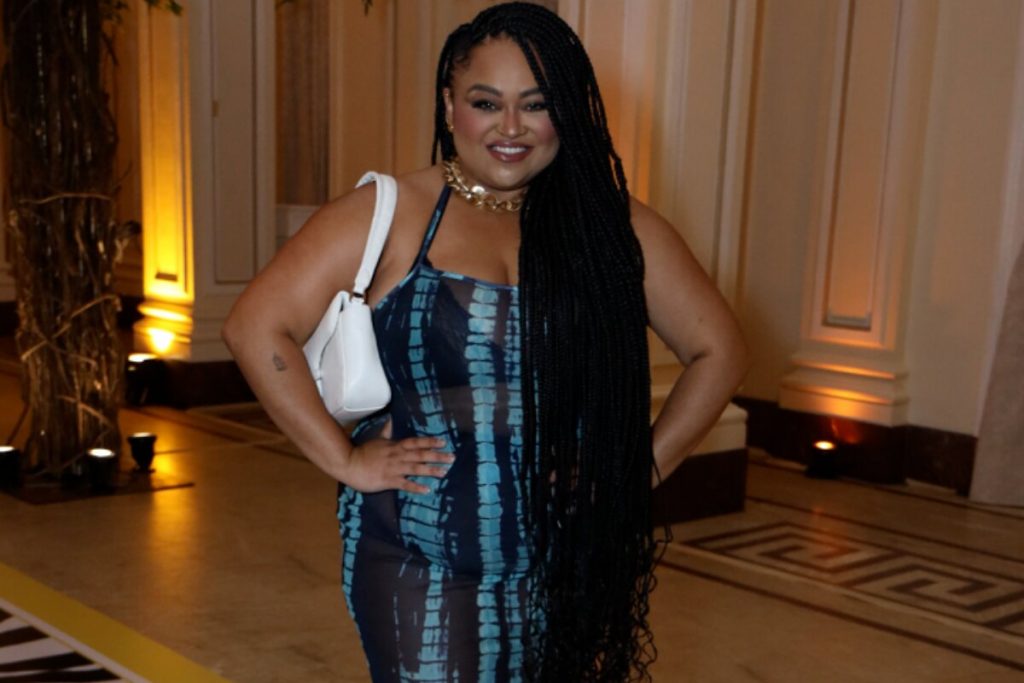 Rízia Cerqueira sorrindo, de mãos na cintura, tranças nos cabelos, vestido azul e preto