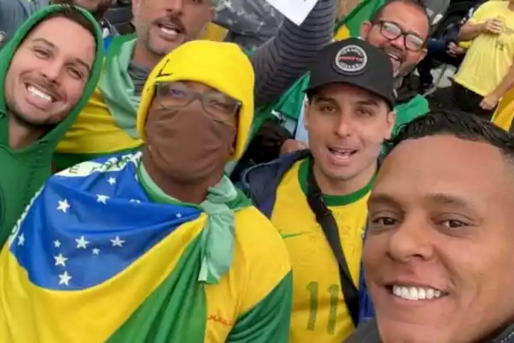 Jogador Robinho disfarçado em manifestação bolsonarista, de gorro amarelo, máscara marrom, coberto com a bandeira do Brasil