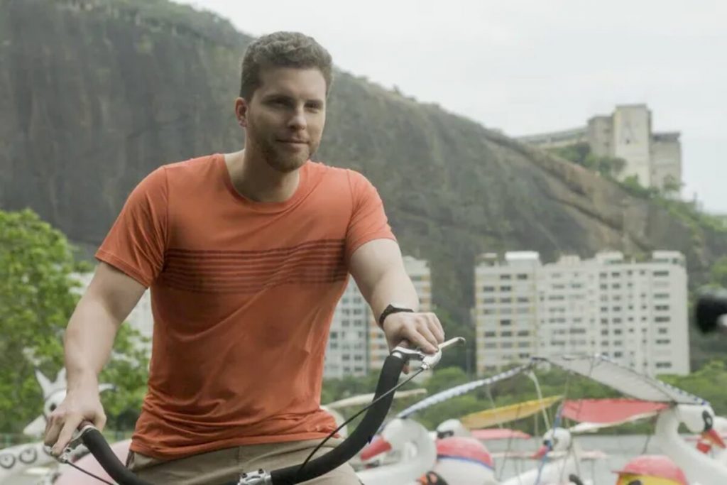 Thiago Fragoso de camisa coral, andando de bike, em "Travessia"